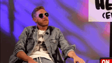 a man wearing sunglasses and a denim jacket is sitting in front of a sign that says on