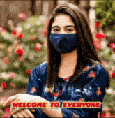 a woman wearing a blue face mask is standing in front of flowers and says welcome to everyone .