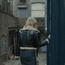 a woman wearing a plaid skirt and a leather jacket stands in an alley