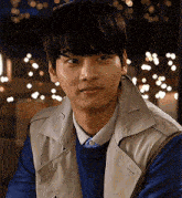 a young man wearing a blue sweater and a tan vest looks at the camera