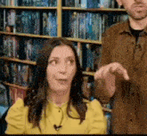 a man and a woman are sitting in front of a bookshelf in a library . the woman is wearing a yellow shirt .