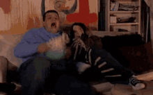 a man and a woman are sitting on a couch eating popcorn and watching a movie .