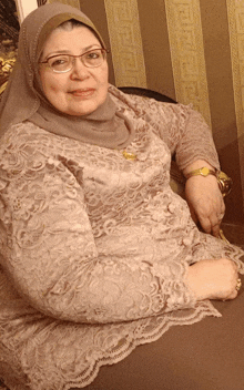a woman wearing glasses and a hijab sits in front of a wall with a greek key pattern