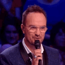 a man wearing glasses and a suit is holding a microphone in front of a crowd .