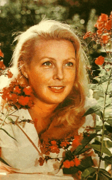 a woman in a white shirt stands in front of a bush with red flowers