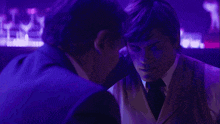 a man in a suit is sitting in front of a bar with purple lights behind him