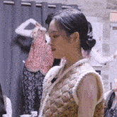 a woman is standing in a room wearing a quilted vest and a bun .