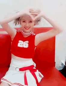 a woman in a cheerleader outfit is sitting on a red couch making a heart with her hands .