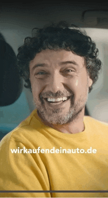 a man with curly hair and a beard is smiling and wearing a yellow shirt with wirkaufendeinauto.de written on it