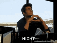 a man is sitting in front of a window making a heart shape with his hands and the words night written below him .