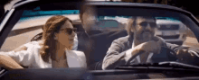 a man and a woman are driving a convertible car . the woman is wearing sunglasses .