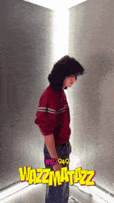 a man in a red sweater is standing in front of a wall that says wazzmatizz