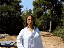 a woman wearing glasses and a white lab coat stands in a field with trees in the background
