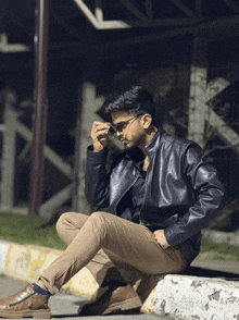a man in a leather jacket sits on a curb with his legs crossed