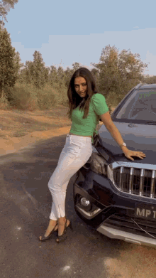 a woman in a green top and white pants leans against a black car with a license plate that says mh
