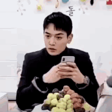 a man is sitting at a table with a bowl of grapes and looking at his phone .