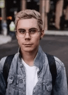 a young man wearing glasses and a denim jacket is standing on the street .