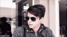 a young man wearing sunglasses and a black jacket is standing in front of a window .