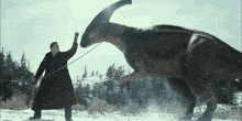 a man in a trench coat stands next to a large dinosaur in the snow