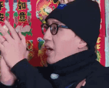 a man wearing glasses and a black hat is clapping his hands in front of a wall with chinese writing on it .