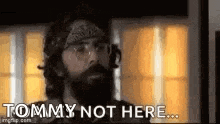 a man with a beard and bandana is standing in front of a window and says `` tommy not here '' .