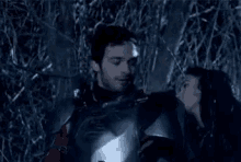 a man in armor is standing next to another man in a dark forest .