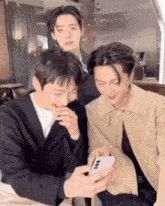 three men are sitting next to each other and one of them is looking at a cell phone .