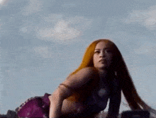 a woman with long red hair is kneeling down on the ground in front of a blue sky .