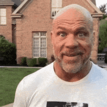 a bald man with a beard wearing a white t-shirt is standing in front of a brick house