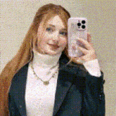 a woman is taking a selfie with her phone while wearing a black jacket and a white turtleneck .