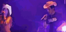 a man and a woman are singing into microphones on a stage . the woman is wearing a cowboy hat .