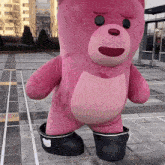 a large pink teddy bear wearing black boots is standing on a sidewalk