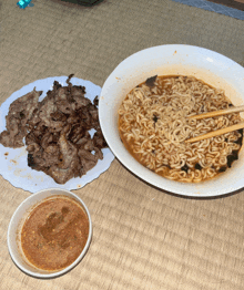 a bowl of ramen next to a plate of meat with chopsticks