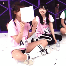 a girl in a pink shirt with the letter a on it holds a piece of paper in front of her face