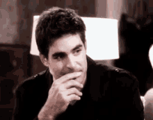 a man in a black jacket is sitting at a table with his hand to his mouth .