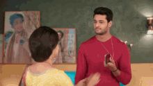 a man in a red shirt is holding a cell phone while talking to a woman in a yellow dress .
