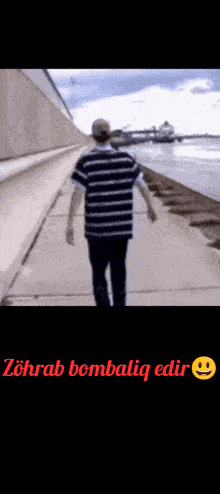 a man in a striped shirt is walking down a sidewalk next to the water .
