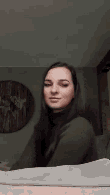 a woman in a green sweater is sitting on a bed in front of a wall clock .