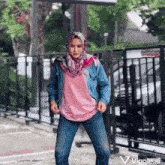 a woman wearing a hijab and a denim jacket is dancing in a parking lot .