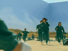a group of people in green uniforms are running on a sandy field .