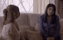 two women are sitting on a couch talking to each other and looking at each other .