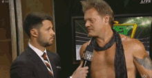 a man in a suit and tie is talking to a shirtless wrestler with a microphone .