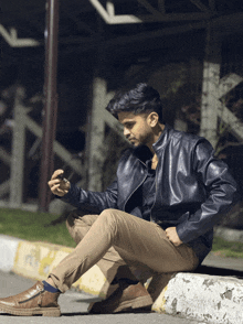a man in a black leather jacket sits on a curb looking at his phone