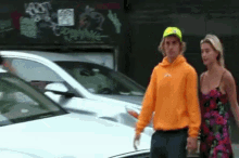 a man in an orange hoodie and a woman in a red dress are walking down a street .