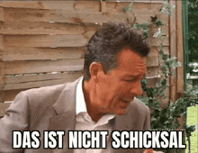 a man in a suit is sitting in front of a wooden fence and says das ist nicht schicksal