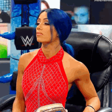 a woman with blue hair and a red dress is sitting in a chair with a w logo in the background