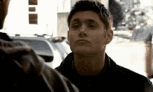 a man in a black jacket is looking at another man in a parking lot .