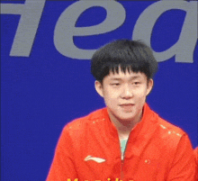 a young man in a red jacket is sitting in front of a blue background with the word tea on it .
