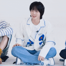 a boy wearing a white shirt with blue flowers on it sits on the floor with his legs crossed