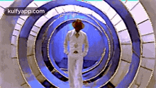 a man in a white suit and hat is walking through a blue spiral tunnel .